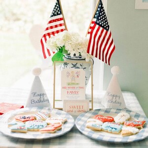 Pode incluir: Uma festa de anivers&aacute;rio com tema vermelho, branco e azul com uma mesa posta com uma toalha de mesa quadriculada azul e branca. H&aacute; dois pratos de biscoitos decorados com glac&ecirc; vermelho, branco e azul. Os biscoitos t&ecirc;m o formato de cora&ccedil;&otilde;es, estrelas e bandeiras. H&aacute; dois chap&eacute;us de festa com as palavras "Feliz anivers&aacute;rio" bordadas. H&aacute; uma placa emoldurada que diz "Red White and Two Hayes' Second Birthday" e uma placa menor que diz "Sweet Land of Liberty".