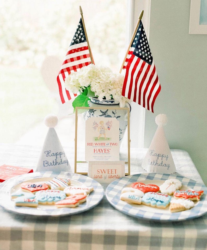 Pode incluir: Uma festa de anivers&aacute;rio com tema vermelho, branco e azul com uma mesa posta com uma toalha de mesa quadriculada azul e branca. H&aacute; dois pratos de biscoitos decorados com glac&ecirc; vermelho, branco e azul. Os biscoitos t&ecirc;m o formato de cora&ccedil;&otilde;es, estrelas e bandeiras. H&aacute; dois chap&eacute;us de festa com as palavras "Feliz anivers&aacute;rio" bordadas. H&aacute; uma placa emoldurada que diz "Red White and Two Hayes' Second Birthday" e uma placa menor que diz "Sweet Land of Liberty".