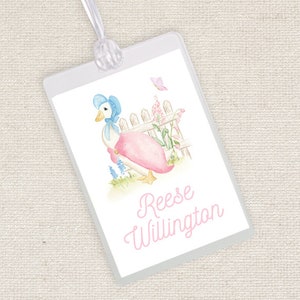 May include: A white goose wearing a blue bonnet and a pink dress stands by a white picket fence with pink flowers. The text "Reese Willington" is written in pink cursive below the goose.