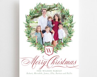 May include: A Christmas card featuring a family portrait framed by a green wreath with red berries. The wreath has a gold and red shield with the letter "W" in the centre. The text "Merry Christmas" is written in red cursive script. The text "The Wilson Family" is written in black below the wreath. The names "Robert, Meredith, James, Ella, Barton and Hollis" are written in black below the family name.