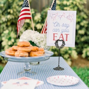 Pode incluir: Uma toalha de mesa quadriculada azul e branca com um prato de donuts, uma placa que diz "Oh say can you EAT", e duas bandeiras americanas.