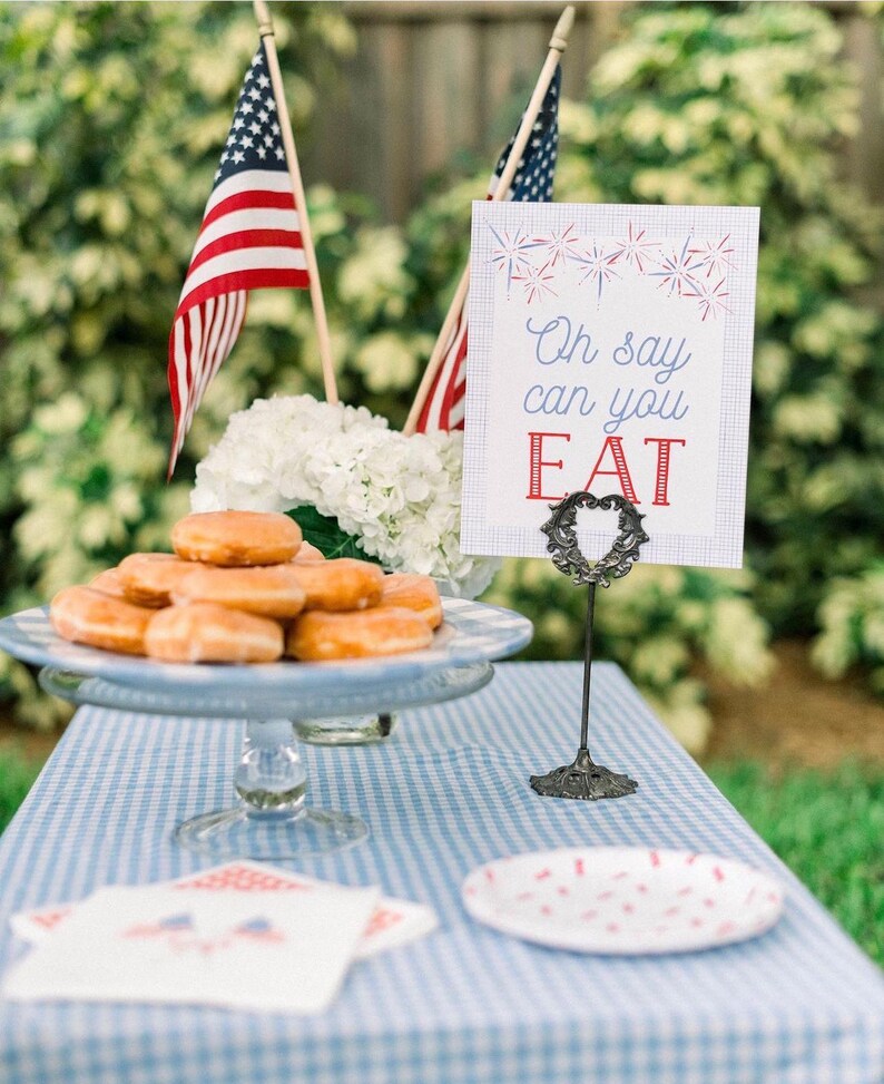 Pode incluir: Uma toalha de mesa quadriculada azul e branca com um prato de donuts, uma placa que diz "Oh say can you EAT", e duas bandeiras americanas.