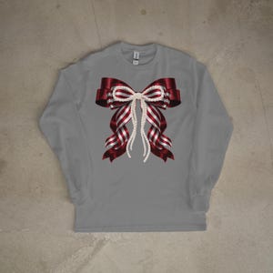 Op de afbeelding: Een grijze longsleeve met een groot strikdesign. De strik heeft een rood-wit gestreept patroon met een parelachtig midden en hangende strengen. Het shirt is een casual stijl.