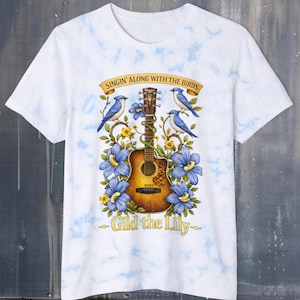 May include: White t-shirt with a watercolour design featuring a brown acoustic guitar, bluebirds, and blue flowers. The text on the shirt reads "Singin' Along With The Birds" and "Gild the Lily".