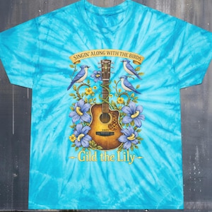 May include: A turquoise tie-dye t-shirt featuring a guitar illustration, blue birds, and flowers. The text reads "Singin' Along With The Birds" and "Gild the Lily". The shirt has a swirling pattern.