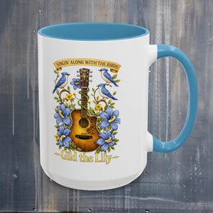 May include: White ceramic mug with a blue interior and handle. The mug features a guitar design with bluebirds, blue flowers, and the text "Singin' Along With The Birds" and "Gild the Lily".