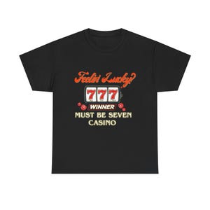 Peut inclure: T-shirt noir avec un motif sur le thème du casino. Le motif présente le texte "Feelin' Lucky?" en orange, une machine à sous avec "777" dans les fenêtres et les mots "Winner" et "Must Be Seven Casino". Deux dés rouges sont également inclus.