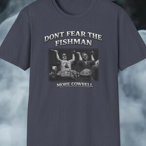 以下が含まれることがあります： 「DON'T FEAR THE FISHMAN」の文字がプリントされた濃い青色のTシャツ。2人の男性の白黒写真が上にあり、その下に「MORE COWBELL」の文字があります。クルーネックのTシャツです。