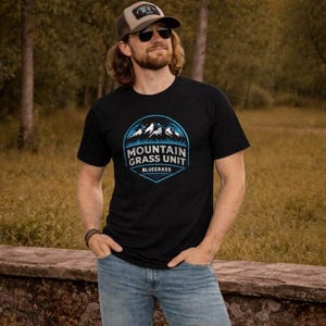 May include: Black t-shirt featuring a blue and white graphic with the text "MOUNTAIN GRASS UNIT BLUEGRASS". The shirt has a crew neck and short sleeves. The person is wearing a baseball cap and sunglasses.