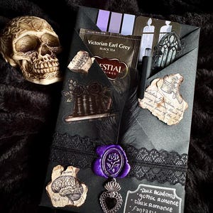 May include: A gothic-themed gift set on a black surface. The set includes a decorative skull, a black envelope with stickers, a tea bag, a feather, a purple wax seal, and a silver heart pendant. The envelope has the words "Dark Academia, gothic Romance, Dark Romance, Fantasy, erotica."