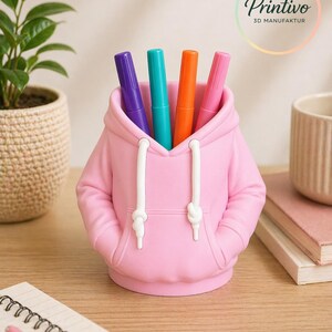 May include: A pink pen holder shaped like a hoodie, with white drawstrings and pockets. The pen holder contains four colorful markers: purple, teal, orange, and pink. The Printivo logo is visible in the background.