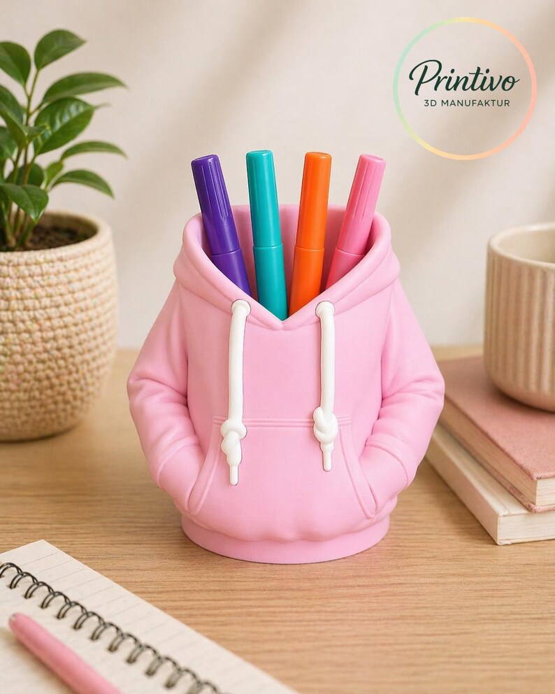 May include: A pink pen holder shaped like a hoodie, with white drawstrings and pockets. The pen holder contains four colorful markers: purple, teal, orange, and pink. The Printivo logo is visible in the background.
