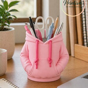 May include: A pink desk organizer shaped like a hoodie, with a hood and pockets. The organizer holds pens, pencils, and scissors. The drawstring is a darker pink. The organizer is on a wooden desk with a laptop and a plant in the background.