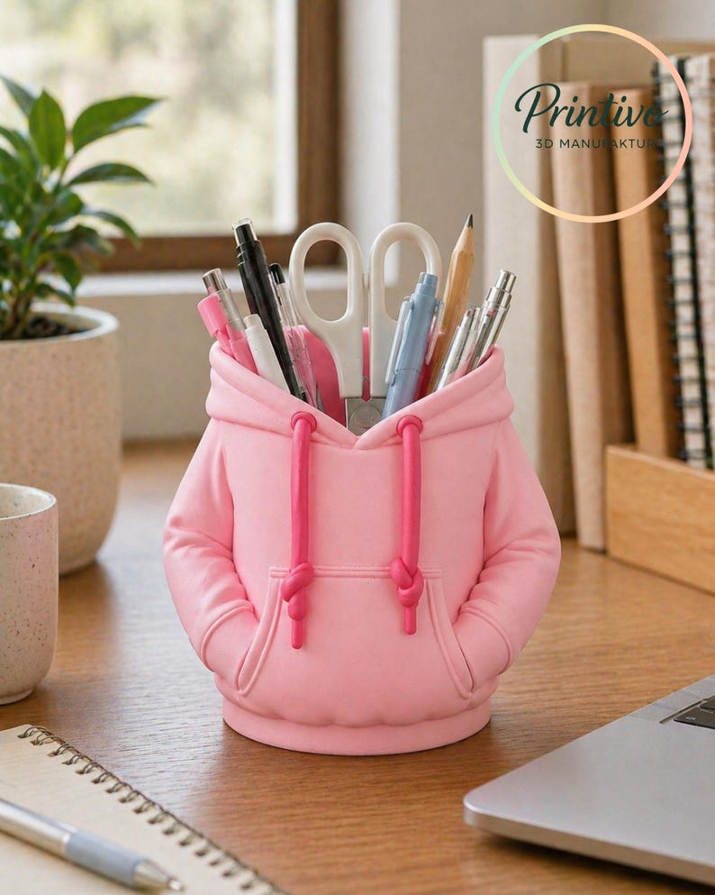 May include: A pink desk organizer shaped like a hoodie, with a hood and pockets. The organizer holds pens, pencils, and scissors. The drawstring is a darker pink. The organizer is on a wooden desk with a laptop and a plant in the background.