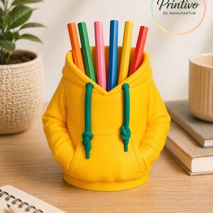 May include: A yellow pencil holder shaped like a hoodie, with green drawstrings and a front pocket. The holder contains several colored pencils in orange, green, pink, blue, yellow, and red. The Printivo logo is visible in the background.