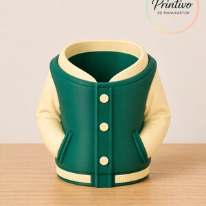 May include: A green and cream-colored varsity jacket-shaped container. The container has a green body with cream-colored sleeves and collar. It features three white buttons down the front and a cream-colored band at the bottom. The container is on a wooden surface.