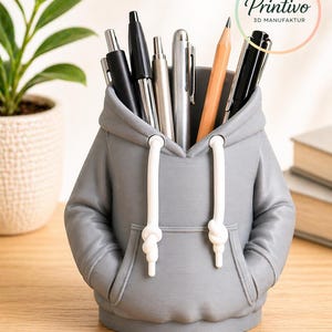May include: Gray pen holder shaped like a hoodie, holding pens and a pencil. The hoodie has white drawstrings and a front pocket. The pen holder is on a wooden surface. The logo "Printivo 3D Manufaktur" is visible.
