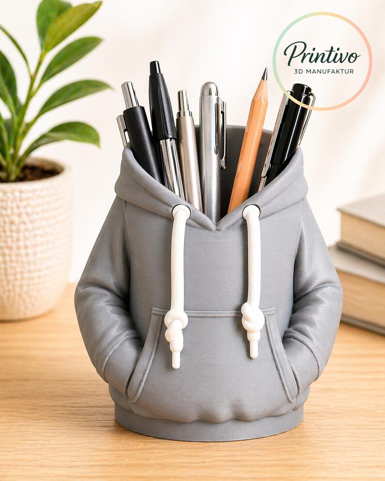 May include: Gray pen holder shaped like a hoodie, holding pens and a pencil. The hoodie has white drawstrings and a front pocket. The pen holder is on a wooden surface. The logo "Printivo 3D Manufaktur" is visible.