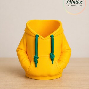May include: A yellow hoodie-shaped container with green drawstrings and pockets. The product is a 3D-printed item. The logo "Printivo 3D MANUFAKTUR" is visible in the upper right corner. The container is sitting on a wooden surface.