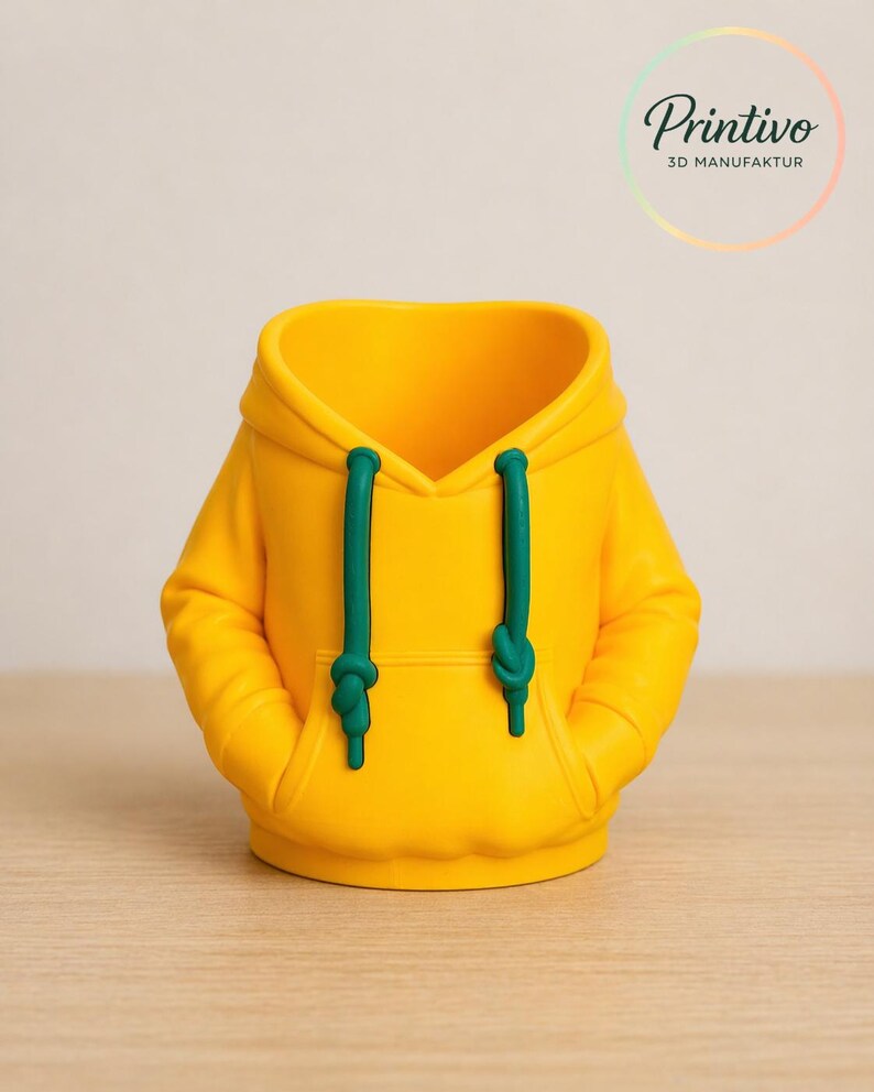 May include: A yellow hoodie-shaped container with green drawstrings and pockets. The product is a 3D-printed item. The logo "Printivo 3D MANUFAKTUR" is visible in the upper right corner. The container is sitting on a wooden surface.