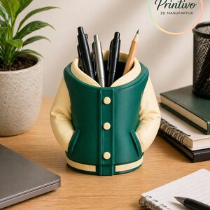 May include: A green and cream-colored desk organizer shaped like a varsity jacket, holding pens and a pencil. The organizer has three buttons and two pockets. The Printivo logo is visible in the background.