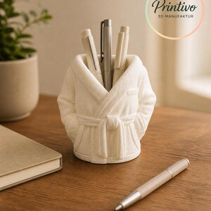 May include: A white pen holder shaped like a bathrobe, holding several pens. The pen holder is on a wooden surface, with a notebook and a pen nearby. The logo "Printivo 3D MANUFAKTUR" is visible in the background.