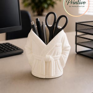 May include: A white robe-shaped desk organizer holding black pens and scissors. The organizer is on a desk with a computer and a plant in the background. The Printivo logo is in the upper right corner.