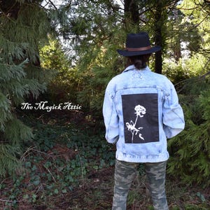 May include: Light blue denim jacket with a black square patch featuring a white rose illustration. The jacket is worn with camouflage pants and a black hat with a brown band. The Magick Attic text is visible in the image.