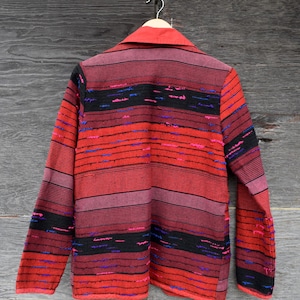 May include: A vintage jacket with a red collar and a striped pattern in shades of red, black, and purple. The jacket is hanging on a wooden hanger against a weathered wooden background. The text "The Magick Attic" is visible.