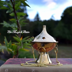 May include: A gold-colored incense burner with a star-shaped cutout on the lid, resting on a vintage book. The burner has a decorative chain and is set against a blurred outdoor background. The text "The Magick Attic" is visible.