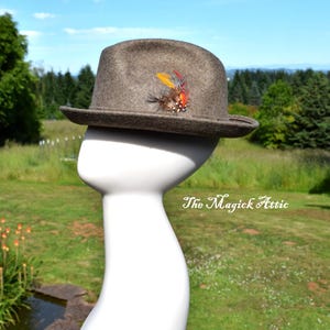 May include: A brown fedora hat with a decorative feather accent. The hat is displayed on a white mannequin head against a backdrop of green grass, trees, and a blue sky. The Magick Attic text is visible.
