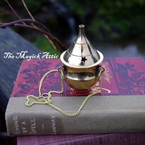 May include: A gold-colored incense burner with a star-shaped cutout on the lid, suspended by a chain. The burner sits atop two antique books with titles including "Bound by a Spell" and "The Strange Woman". The text "The Magick Attic" is visible.
