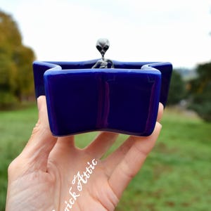 May include: A deep blue, star-shaped ceramic dish with a small, silver alien figurine inside. The dish is held in a hand, with a blurred green background. The Magick Attic is written on the hand.