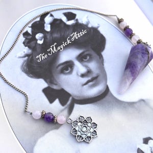 May include: A silver-toned pendulum necklace with a flower charm and a purple amethyst crystal point. The necklace features a chain with pink and purple beads. The image has the text "The Magick Attic" on a vintage photograph.