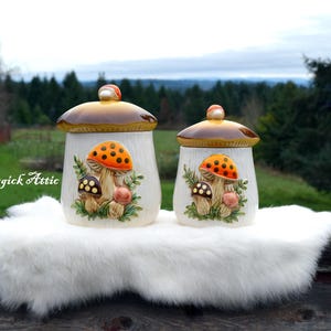 May include: Two ceramic mushroom-themed canisters with lids. The canisters are white with a brown and orange mushroom design. The lids are brown with a small orange knob. The canisters are displayed on a white fluffy surface.