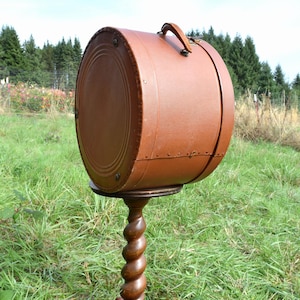 May include: A brown, round vintage hat box with a leather handle, mounted on a wooden stand with a spiral-turned column. The hat box is a warm brown color and sits in a grassy field.