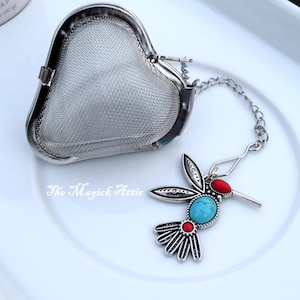 May include: A silver-toned tea infuser shaped like a heart, with a chain attached to a hummingbird charm. The hummingbird has turquoise and red accents. The Magick Attic is printed on the surface.
