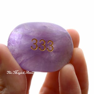 May include: A lavender-colored, oval-shaped stone with the gold number "333" etched on its surface. The stone is held in a person's hand against a white background. The Magick Attic is written on the bottom left.