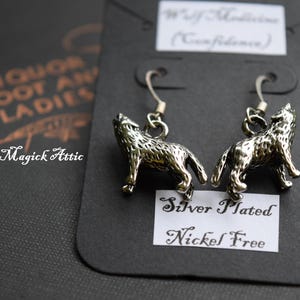 May include: Silver-plated wolf earrings with a howling wolf design. The earrings are nickel-free and are displayed on a black card with the text "Wolf Medicine (Confidence)". The Magick Attic logo is also visible.