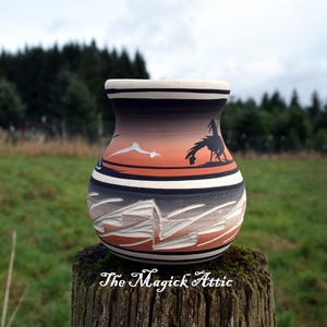 May include: A handcrafted ceramic vase with a unique design. The vase features a gradient of colors, including orange, black, and white, with a detailed landscape and animal silhouette. The Magick Attic is printed on the bottom.