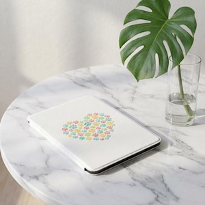 Op de afbeelding: Witte tablethoes met een hartvormig ontwerp van kleurrijke pootafdrukken. De hoes ligt op een marmeren tafel, met een groen blad en een glas water op de achtergrond. De hoes heeft een zwarte rand.