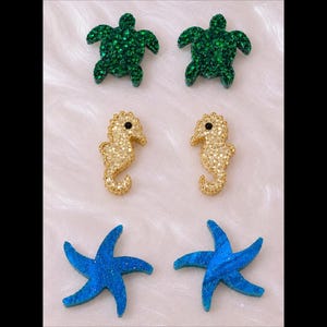 May include: Three pairs of stud earrings featuring sea-themed designs. The top pair consists of green sea turtles with a glittery finish. The middle pair features gold-toned seahorses with small black eyes. The bottom pair shows blue starfish.