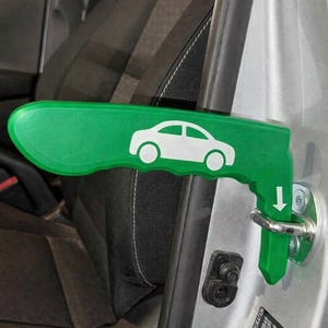 May include: A green metal car door assist handle featuring a white car icon and a downward-pointing arrow. The tool is hooked into the silver metal door latch mechanism of a vehicle, providing a sturdy grip for entry and exit assistance.
