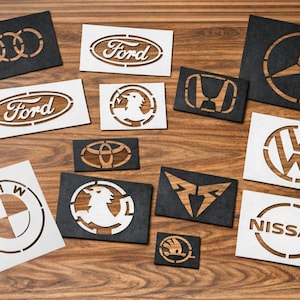 Op de afbeelding: Een verzameling houten logo's van automerken, waaronder Audi, Ford, BMW, Mercedes-Benz, Volkswagen en Nissan. De logo's zijn lasergesneden met een contrasterende houtnerf, op zwarte of witte achtergronden.