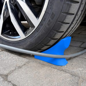 Op de afbeelding: Een felblauwe kunststof kabelbeschermer geplaatst direct achter een zwarte autoband. De oprit heeft een gebogen kanaal dat is ontworpen om een grijze oplaadkabel stevig op zijn plek te houden, zodat deze niet door het wiel wordt geplet.