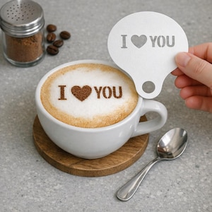 May include: A white ceramic coffee cup with a heart-shaped design and the words "I ❤️ YOU" in the foam. A stencil with the same design is held above the cup. A small spoon and a jar of cinnamon are also present.