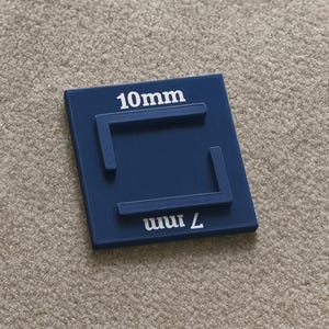 Pode incluir: Um quadrado azul marinho com texto branco indicando 10 mm e 7 mm. O quadrado tem elementos retangulares em relevo. O objeto é provavelmente uma ferramenta de medição ou um calibrador. O fundo é uma superfície texturizada neutra.