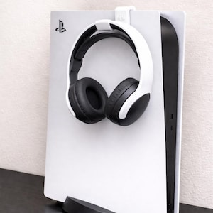 Può includere: Una cuffia da gioco bianca e nera è appoggiata su una console PlayStation 5 bianca. La console è in posizione verticale su un supporto nero. Il logo PlayStation è visibile.