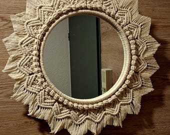 May include: A round mirror with a cream-colored macrame sunburst design. The mirror has a circular reflective surface surrounded by intricate woven patterns and fringed edges, creating a bohemian aesthetic. The mirror is mounted on a wooden surface.