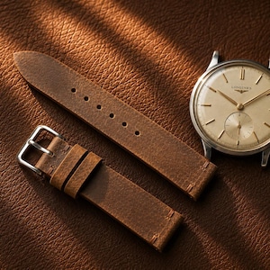 Op de afbeelding: Een zilverkleurig polshorloge met een ronde crèmekleurige wijzerplaat voorzien van het Longines-logo, dunne uurmarkeringen en een kleine sub-wijzerplaat. Het horloge wordt vergezeld door een tweedelige bruine lederen band met een zilveren metalen gesp.
